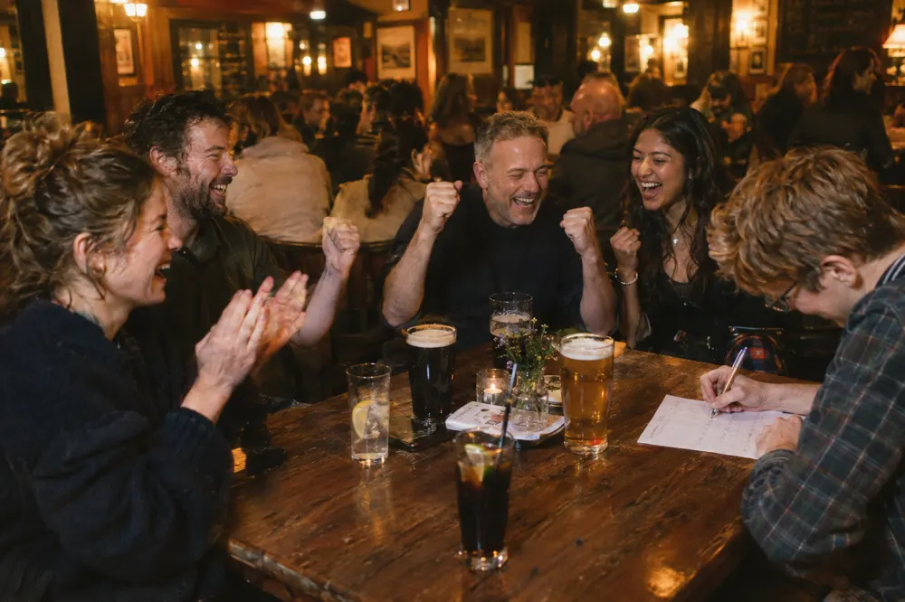 A pub quiz team of five cheering around a wooden table